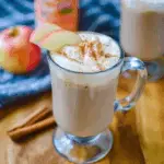 A clear glass mug filled with apple butter latte, topped with frothy whipped cream, a sprinkle of cinnamon, and garnished with fresh apple slices.