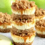 Stack of apple crisp mini cheesecakes with crumb topping and caramel drizzle, surrounded by green apples and apple slices.