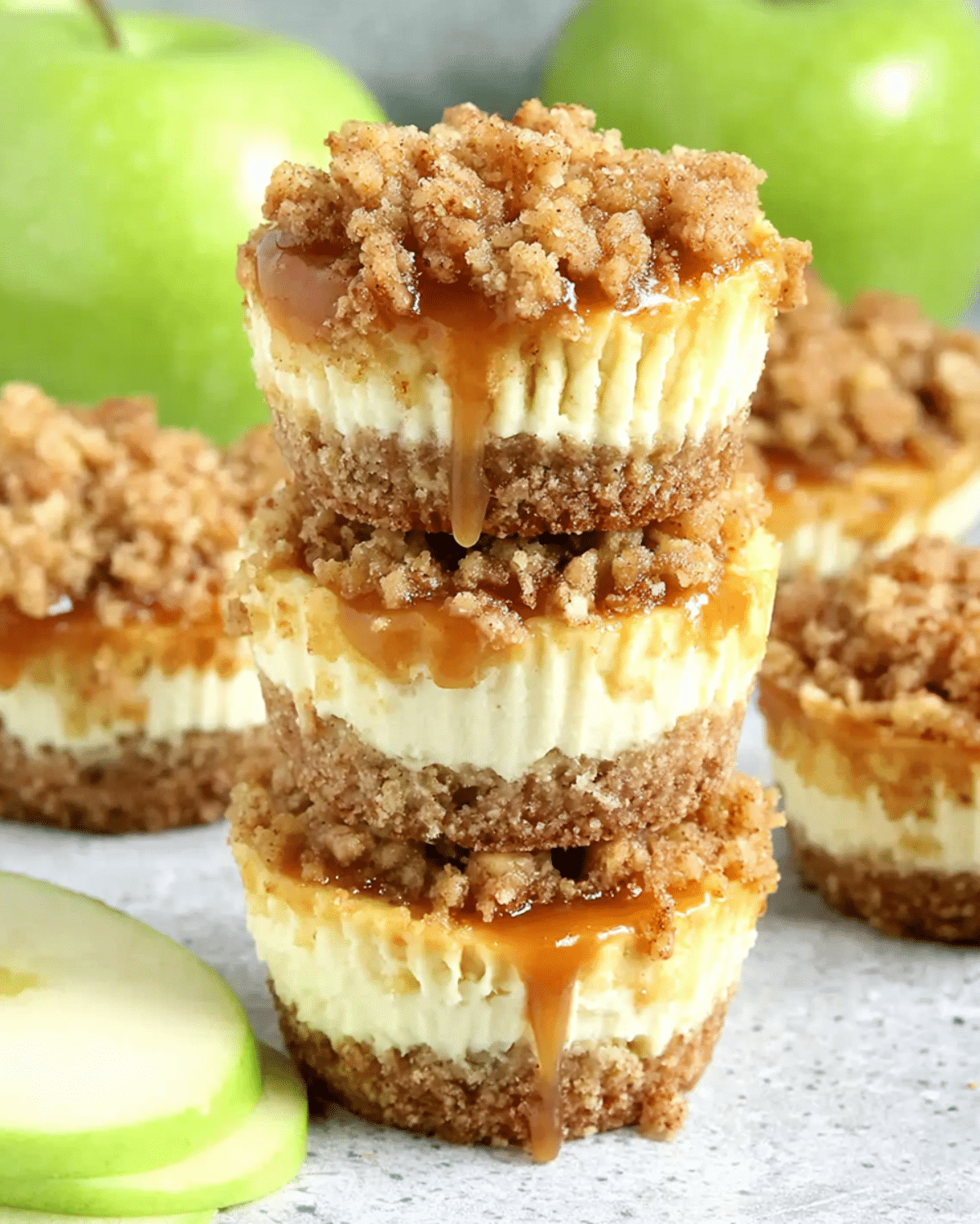 Stack of apple crisp mini cheesecakes with crumb topping and caramel drizzle, surrounded by green apples and apple slices.
