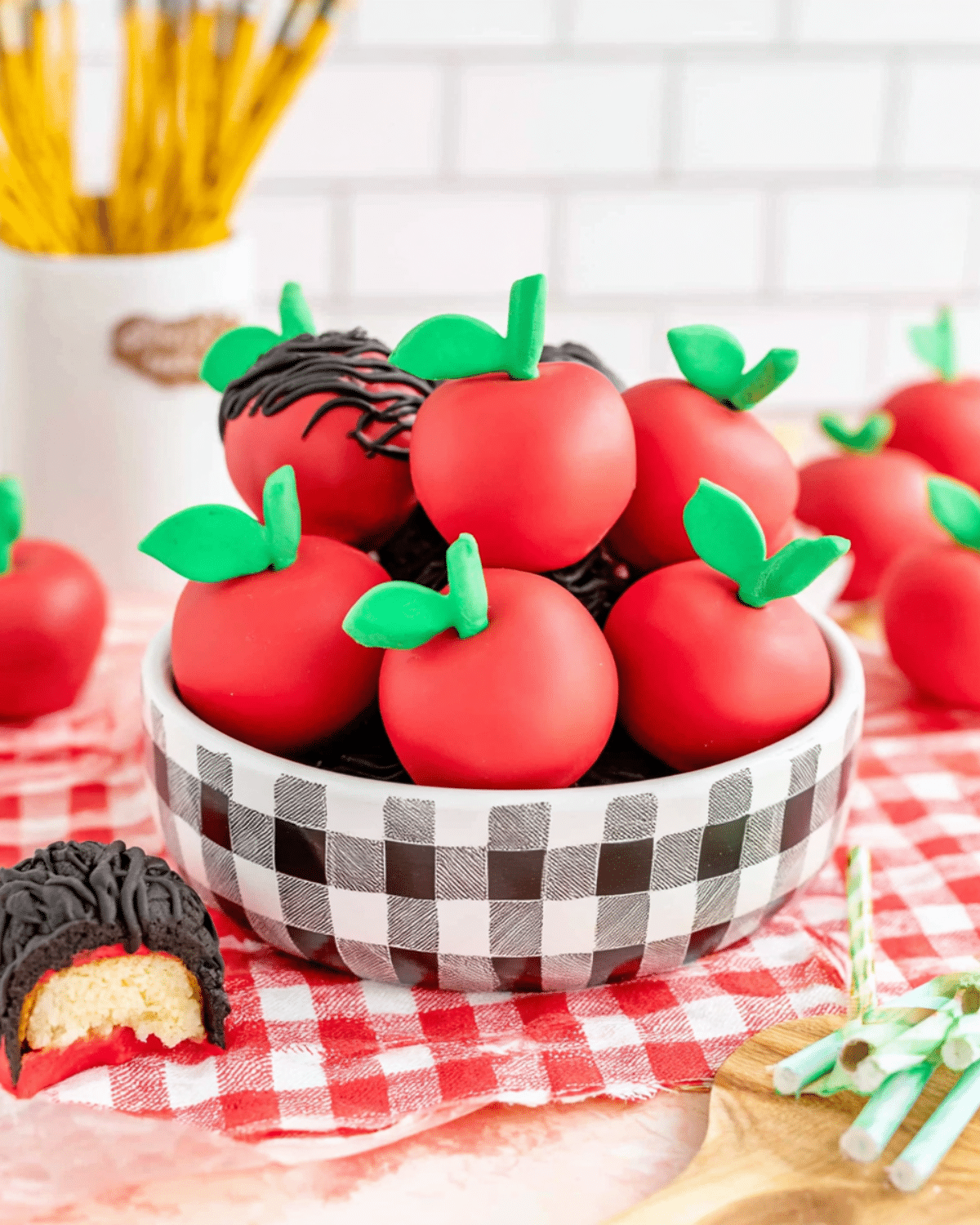 Apple OREO balls decorated in red with green candy leaves and stems, displayed in a checkered bowl on a red gingham cloth.