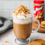 A glass mug of Biscoff cookie butter latte topped with whipped cream, cookie crumbs, and a drizzle of Biscoff spread, with cookies and a jar of Biscoff in the background.
