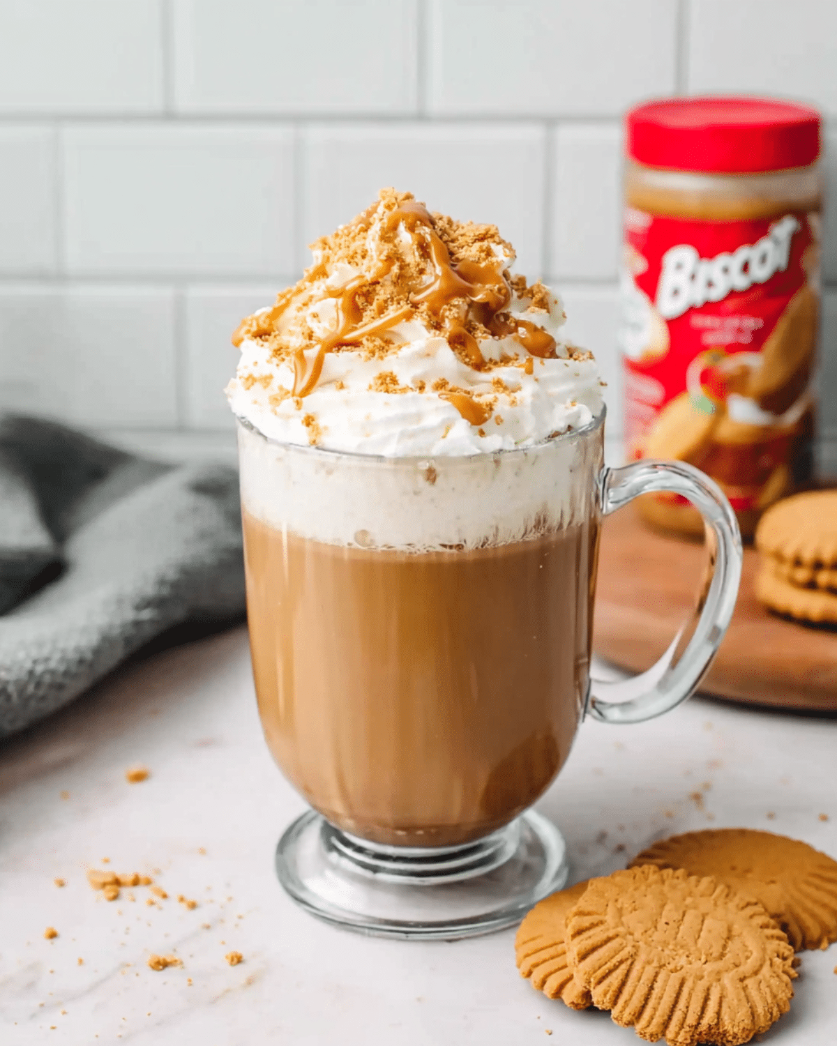 A glass mug of Biscoff cookie butter latte topped with whipped cream, cookie crumbs, and a drizzle of Biscoff spread, with cookies and a jar of Biscoff in the background.