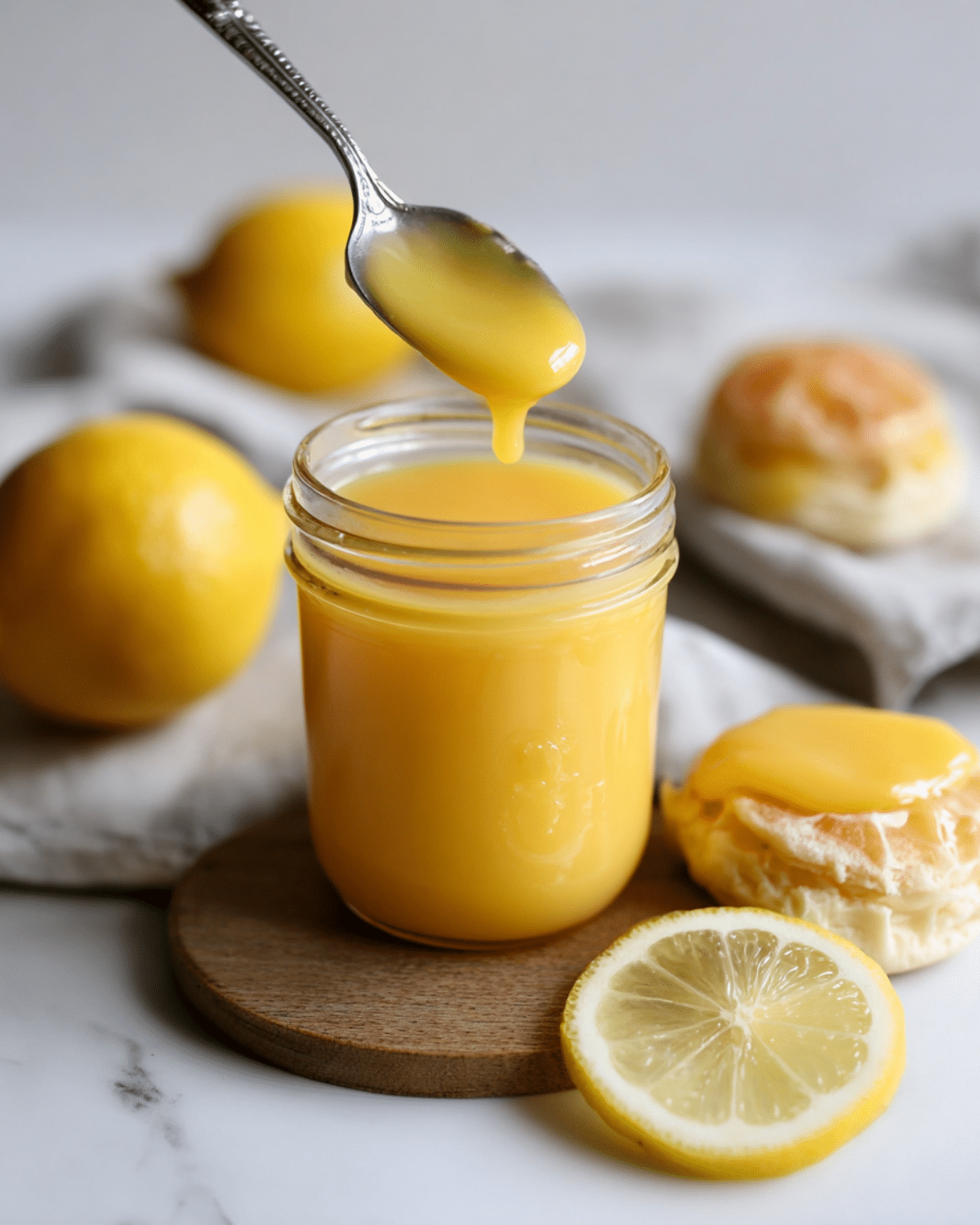 A spoon dripping golden lemon curd into a mason jar, surrounded by fresh lemons, a biscuit with curd, and a lemon slice on a wooden coaster.