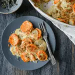 Cheesy Root Vegetable Gratin with layers of sliced sweet potatoes and white potatoes baked in a creamy cheese sauce, served on a dark plate.