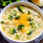 Creamy broccoli cheese soup served in a white bowl, topped with shredded cheddar and surrounded by fresh broccoli and a bread roll.