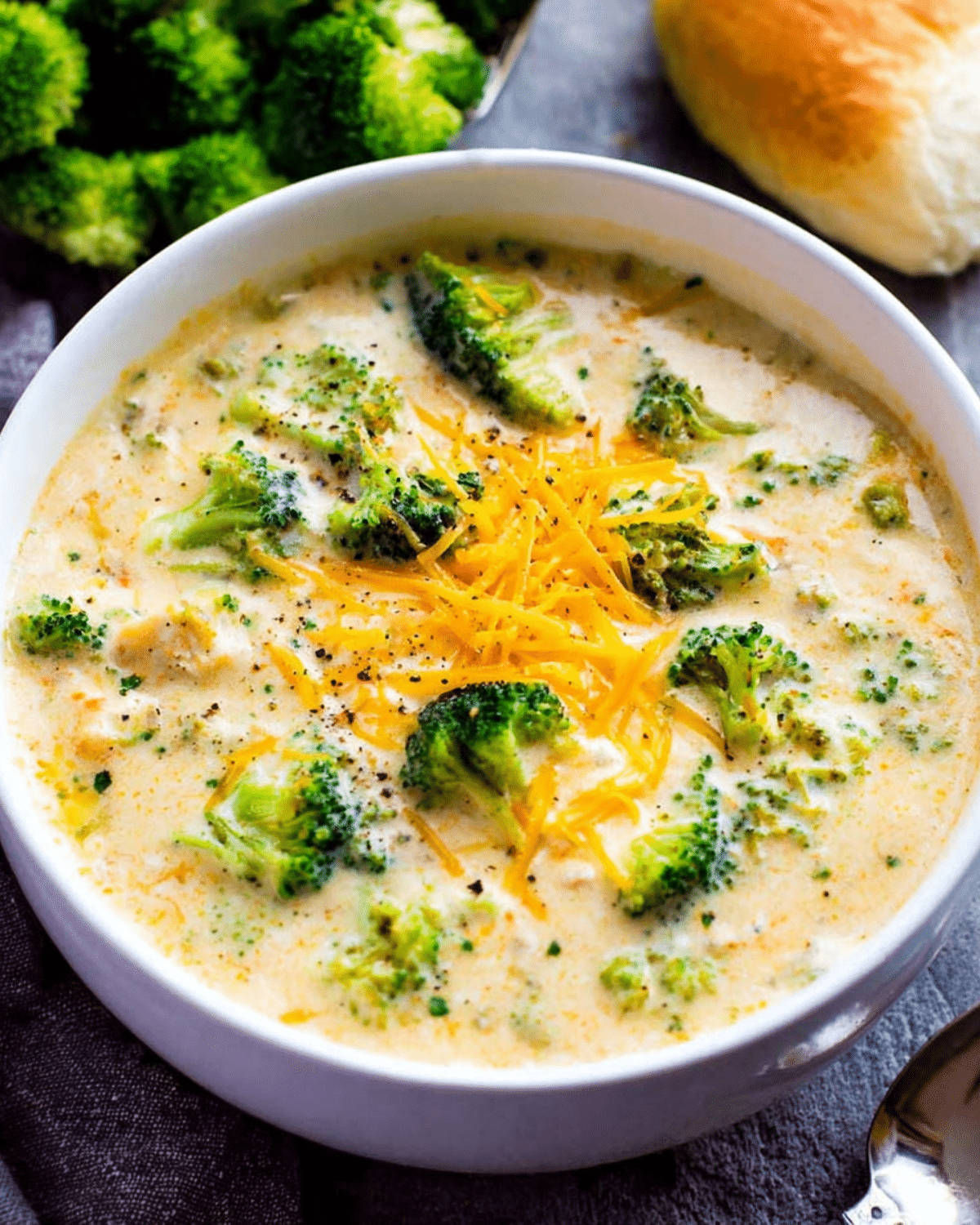 Creamy broccoli cheese soup served in a white bowl, topped with shredded cheddar and surrounded by fresh broccoli and a bread roll.
