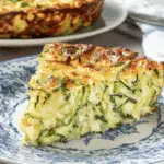A slice of crustless zucchini quiche on a blue and white floral plate, with the full quiche in the background.