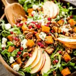 Fall Harvest Salad with roasted butternut squash, apple slices, pecans, goat cheese, pomegranate seeds, and arugula in a wooden bowl.