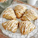 A plate of golden Flaky Apricot Scones drizzled with white icing, arranged neatly on a decorative dish with a soft plaid napkin underneath.