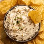 A bowl of creamy French onion dip garnished with chopped chives, surrounded by ridged potato chips, with one chip dipped into the center.