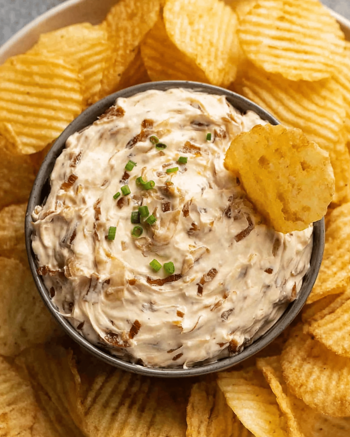 A bowl of creamy French onion dip garnished with chopped chives, surrounded by ridged potato chips, with one chip dipped into the center.