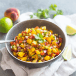 Fresh Peach Salsa in a bowl with diced peaches, tomatoes, red onion, cilantro, and lime wedges on the side.