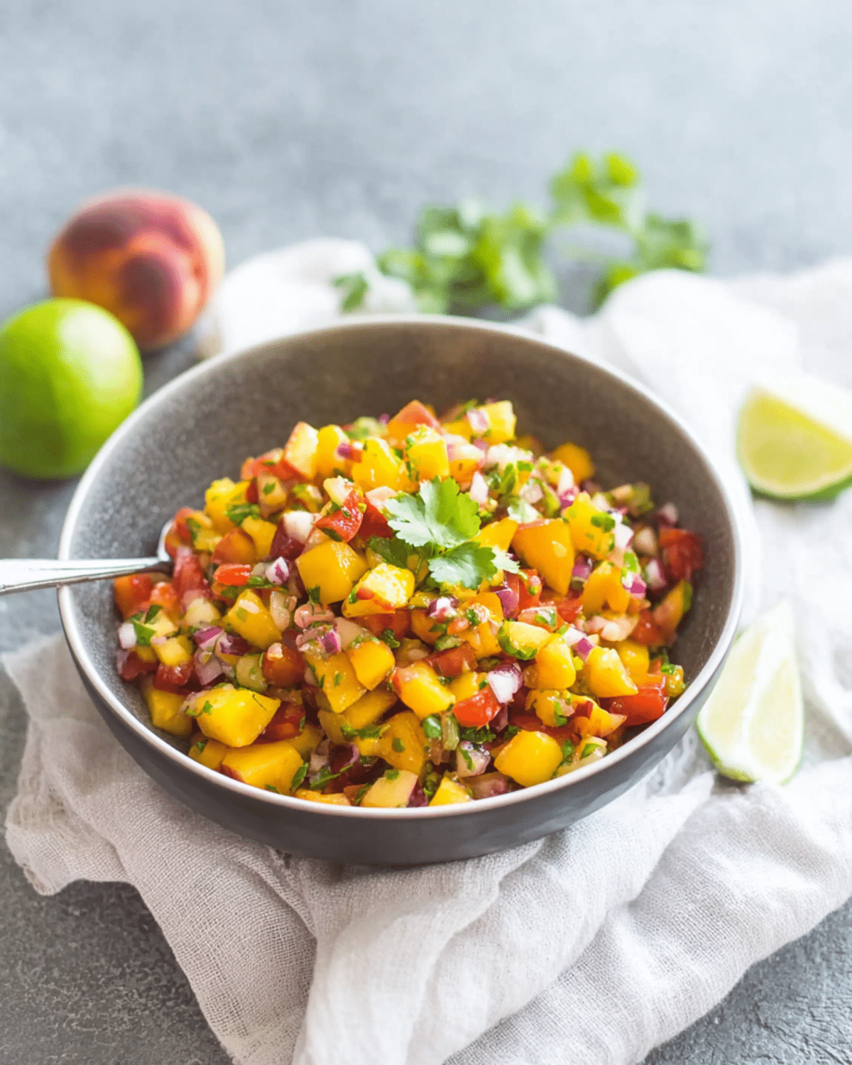 Fresh Peach Salsa in a bowl with diced peaches, tomatoes, red onion, cilantro, and lime wedges on the side.