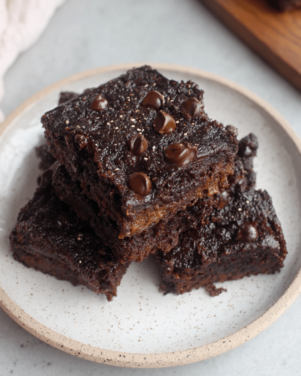 A stack of healthy pumpkin brownies with a rich chocolate texture and chocolate chips on top, served on a ceramic plate.