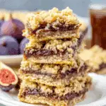 Stack of golden Homemade Fig Bars with a crumbly oat topping and a thick fig filling, served on a white plate with fresh figs in the background.
