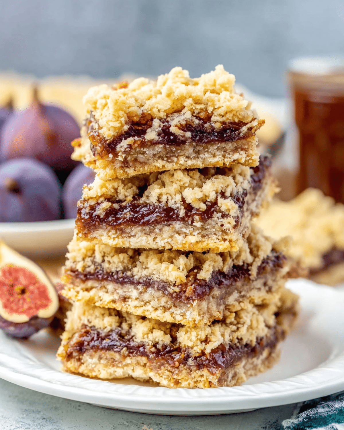 Stack of golden Homemade Fig Bars with a crumbly oat topping and a thick fig filling, served on a white plate with fresh figs in the background.