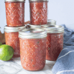 Jars of homemade salsa for canning on a marble countertop with a lime and kitchen towel nearby.