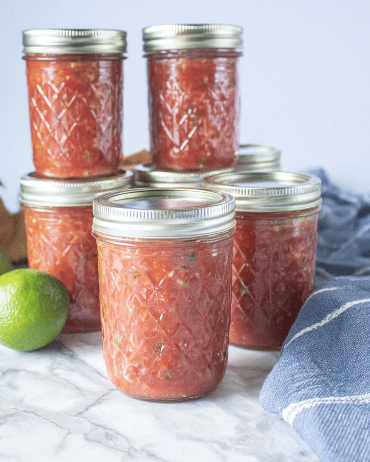 Jars of homemade salsa for canning on a marble countertop with a lime and kitchen towel nearby.