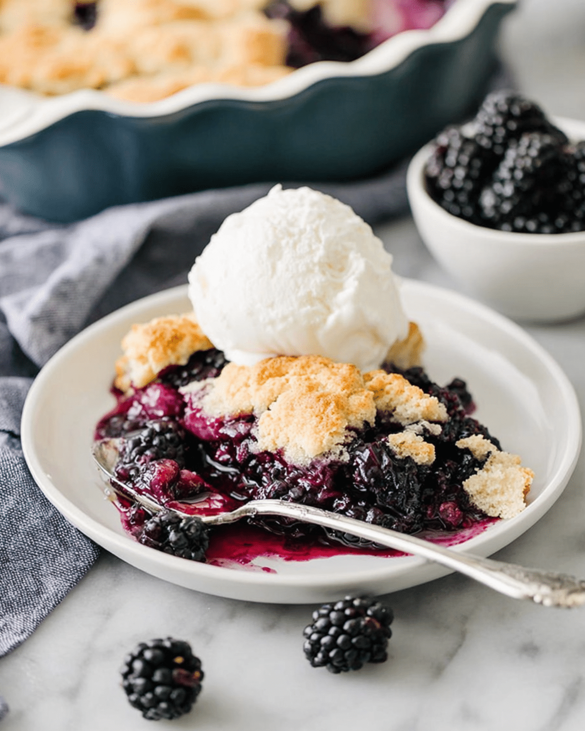 A serving of Kentucky blackberry cobbler topped with vanilla ice cream on a white plate, surrounded by fresh blackberries and a baking dish in the background.