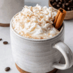 A rustic ceramic mug filled with maple cinnamon latte, topped with whipped cream, sprinkled with cinnamon, and garnished with a cinnamon stick.