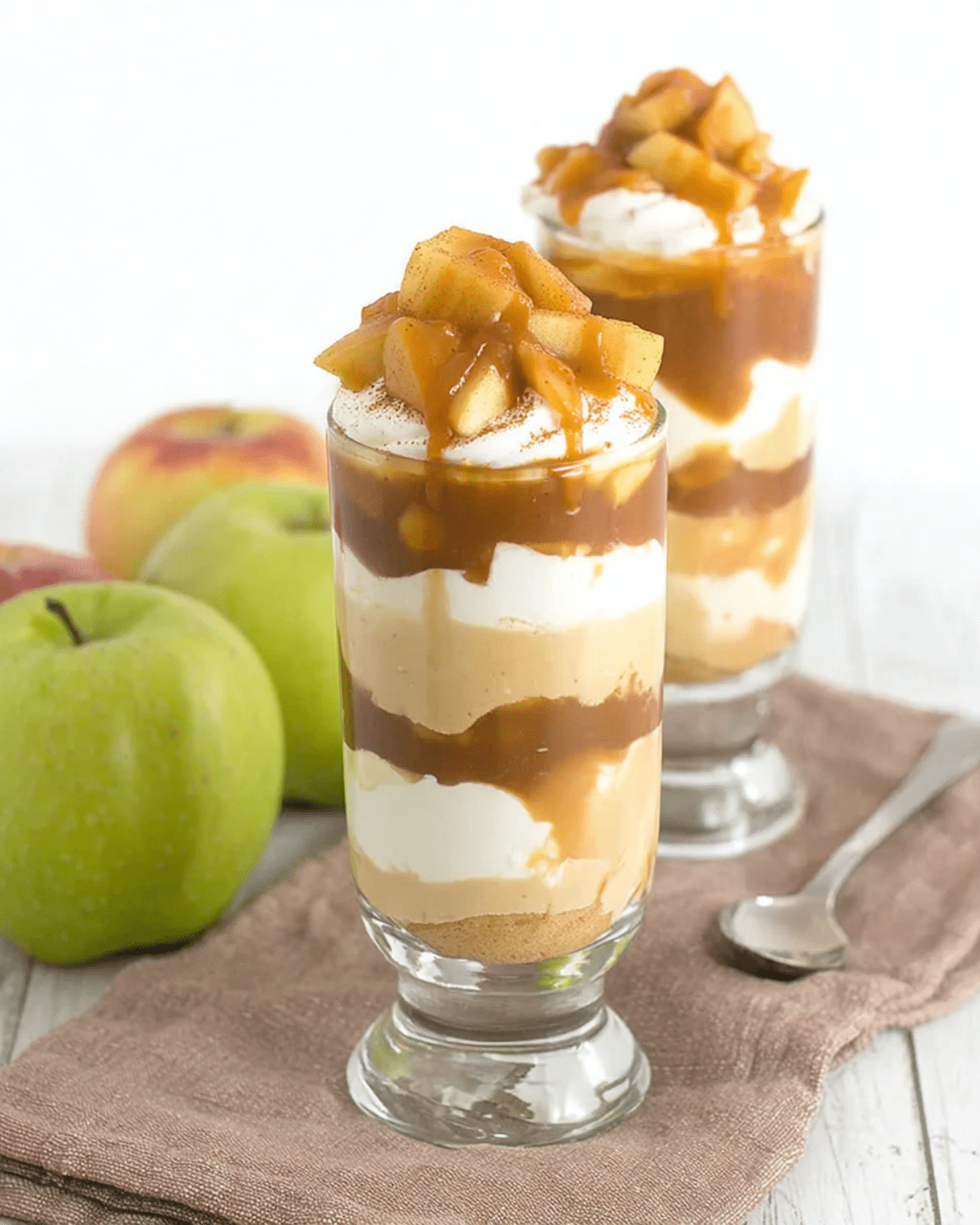 No bake caramel apple pie cheesecake parfaits layered with whipped cream, caramel, and apple topping in tall glasses.