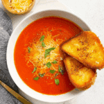 Old-fashioned cream of tomato soup served in a white bowl, topped with shredded cheese and fresh basil, accompanied by two slices of golden grilled bread.