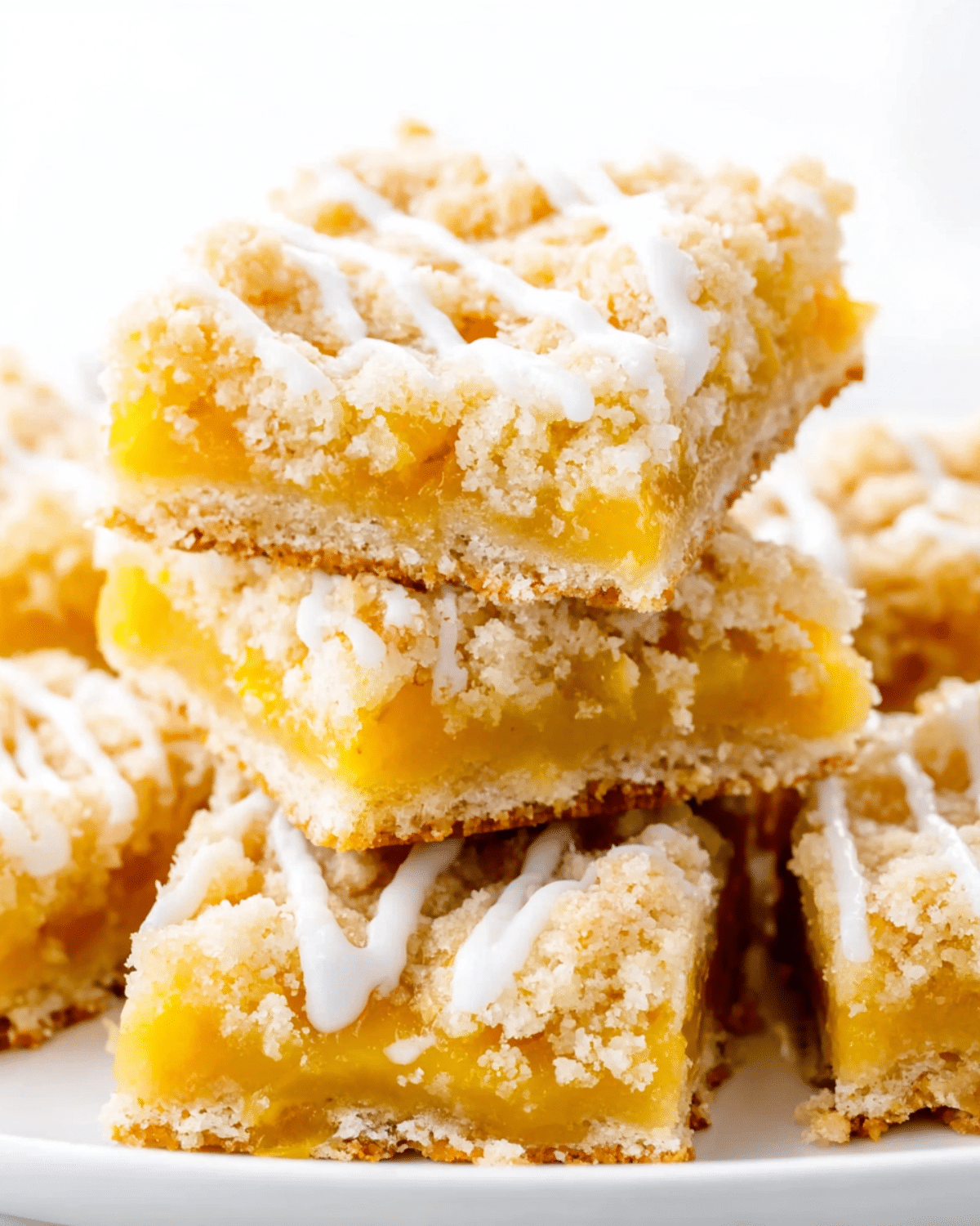 Peach Bars stacked on a white plate, featuring a crumbly topping, sweet peach filling, and a drizzle of white icing.