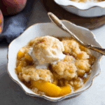 Peach cobbler served in a white scalloped bowl with a scoop of vanilla ice cream on top and a golden spoon.