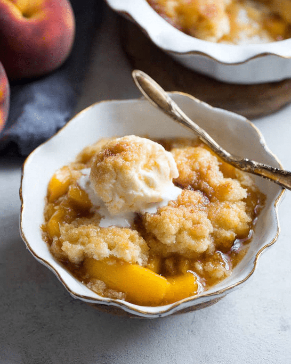 Peach cobbler served in a white scalloped bowl with a scoop of vanilla ice cream on top and a golden spoon.