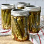 Jars of pickled okra sealed with metal lids, filled with whole okra, garlic, and spices on a wooden surface.