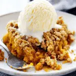 Warm serving of pumpkin crisp topped with a scoop of vanilla ice cream on a white plate with a spoon.