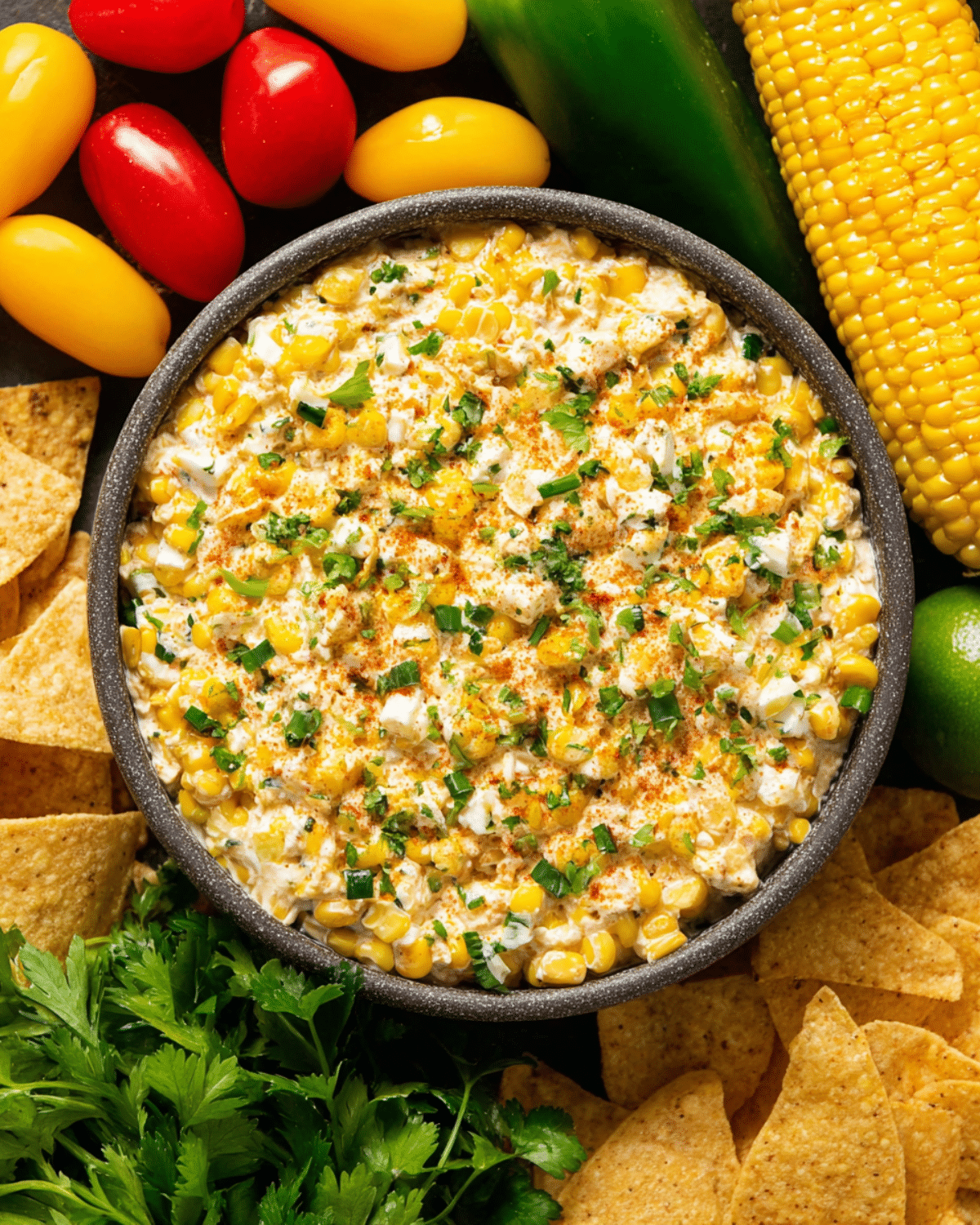 Quick Elote-Inspired Corn Dip served in a bowl, surrounded by fresh tomatoes, corn, tortilla chips, lime, and herbs.