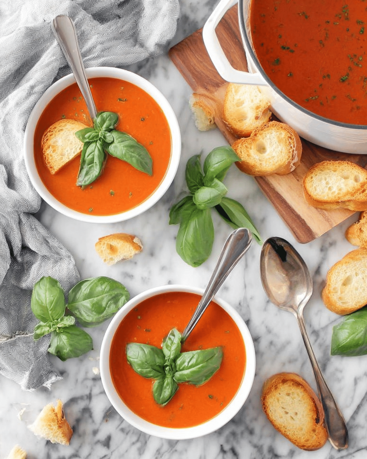Bowls of Roasted Tomato Basil Soup garnished with fresh basil leaves and served with crispy baguette slices on a marble surface.