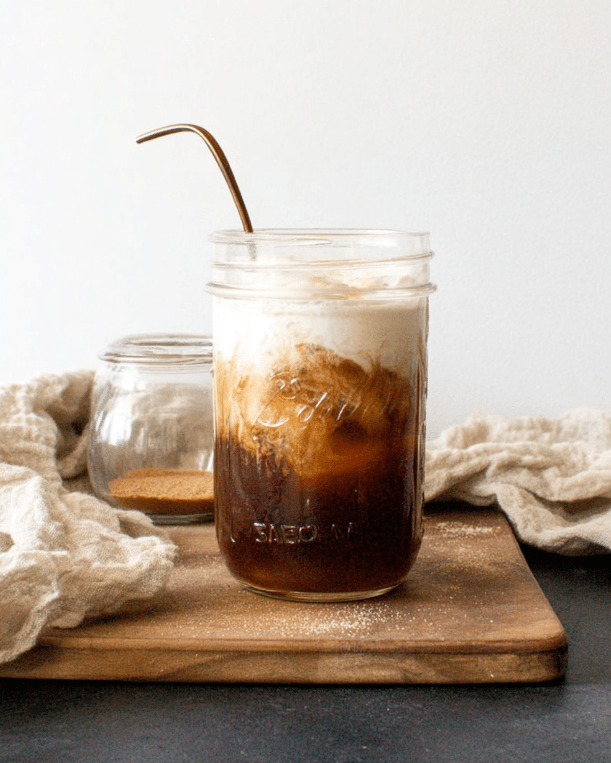 A mason jar filled with salted maple cinnamon cold brew, topped with creamy foam and swirled with milk, served with a metal straw on a wooden board.