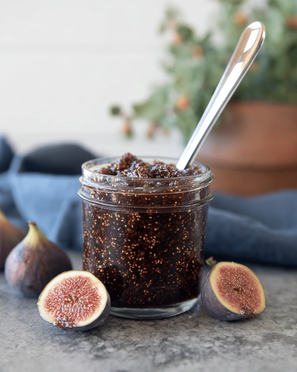 Glass jar filled with Small-Batch Fig Honey Jam, surrounded by fresh figs and a spoon resting inside the jar on a gray countertop.