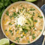 Bowl of White Chicken Chili topped with shredded cheese, avocado, and fresh cilantro, served with lime wedges and avocado on the side.