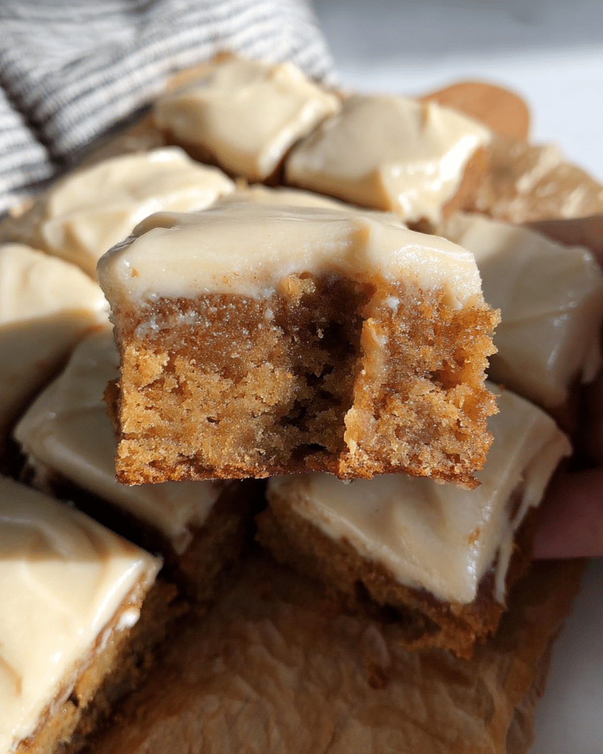 A close-up of a bitten apple cinnamon blondie topped with maple cream cheese frosting, with more blondies in the background.