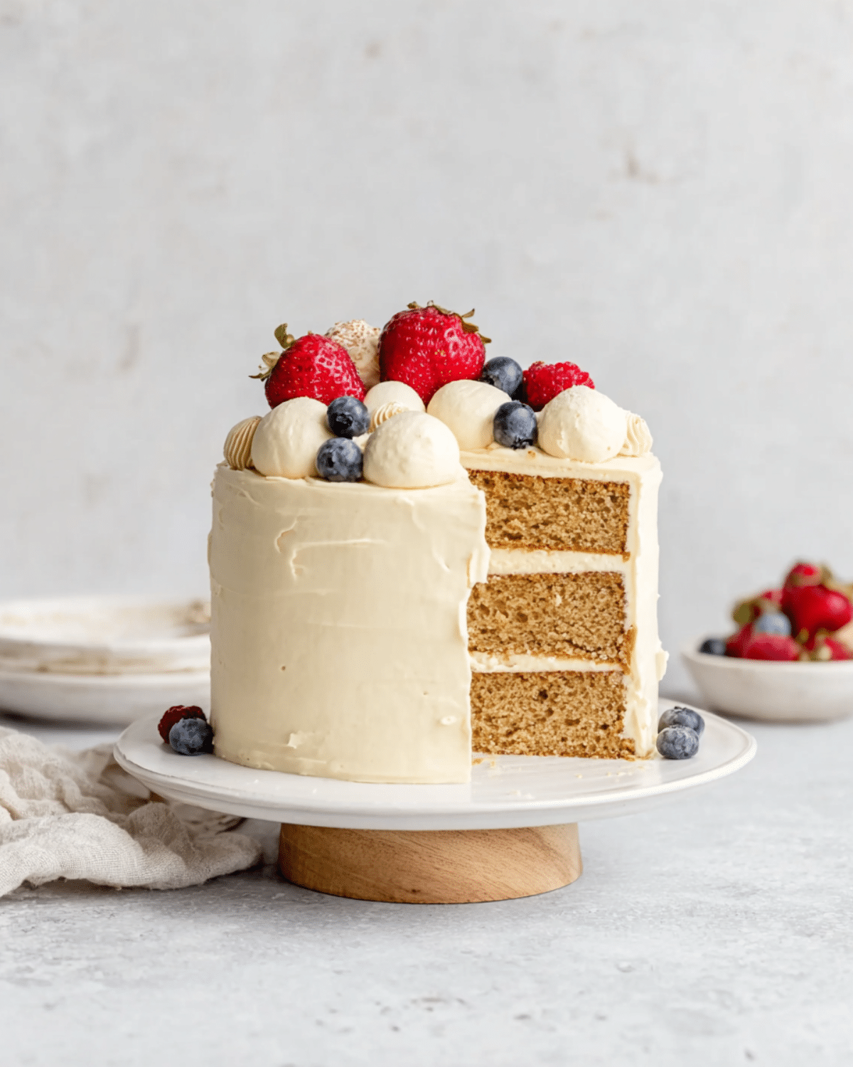 A three-layer baby birthday cake with smooth white frosting, topped with strawberries, blueberries, and piped dollops of cream, with a slice removed to show the interior.