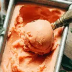 Creamy peach sorbet being scooped from a metal container with a vintage ice cream scooper.