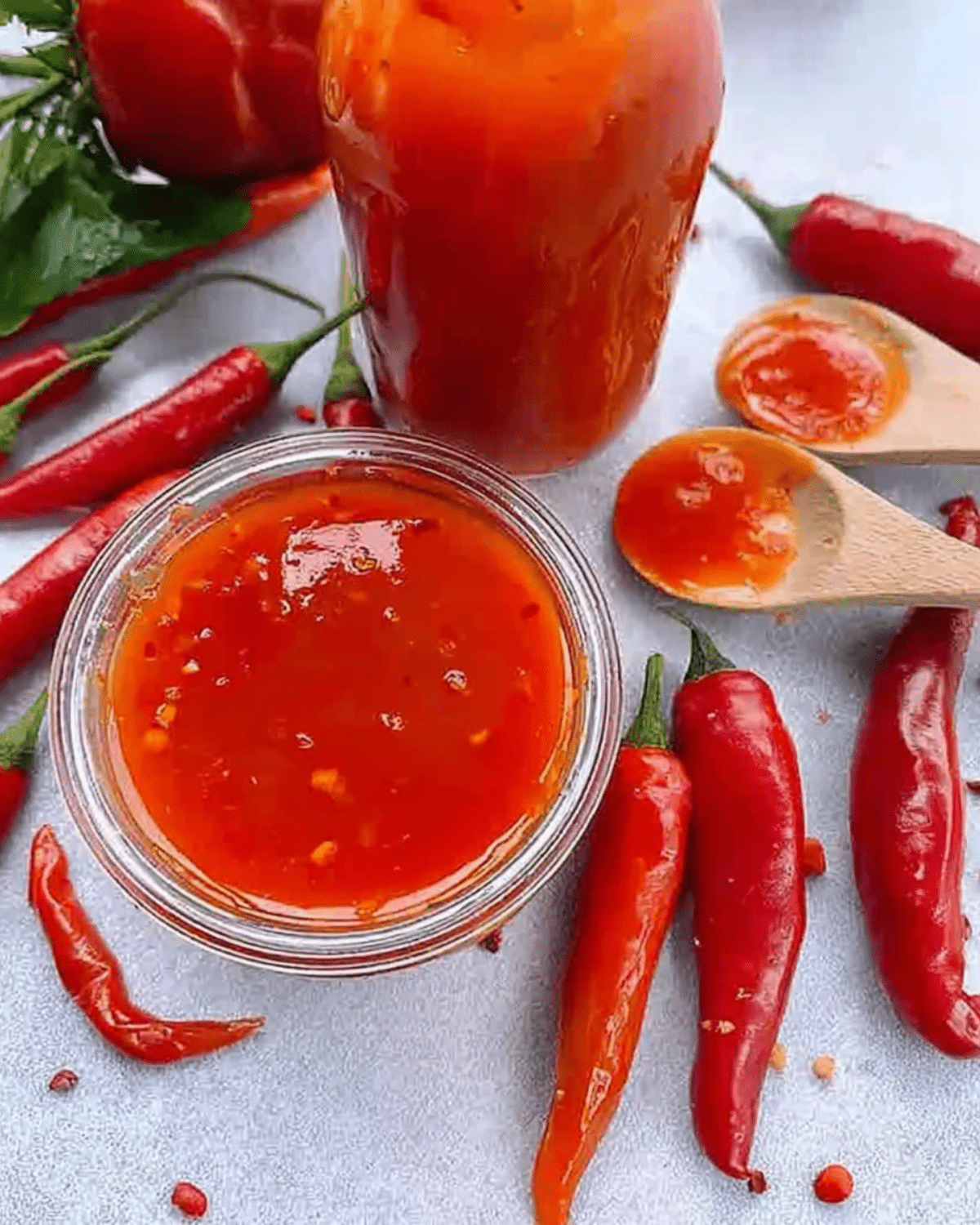 Glass jar and bowl of vibrant red pepper hot sauce surrounded by fresh red chili peppers.