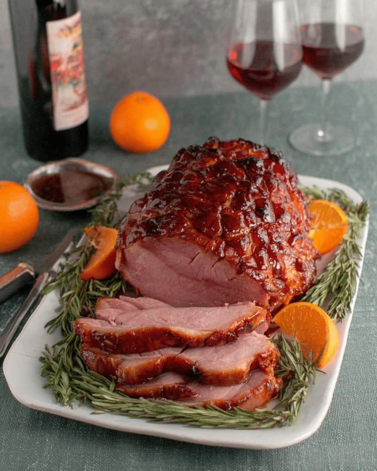 Sliced brown sugar rum baked ham on a white platter, garnished with rosemary sprigs and orange slices.