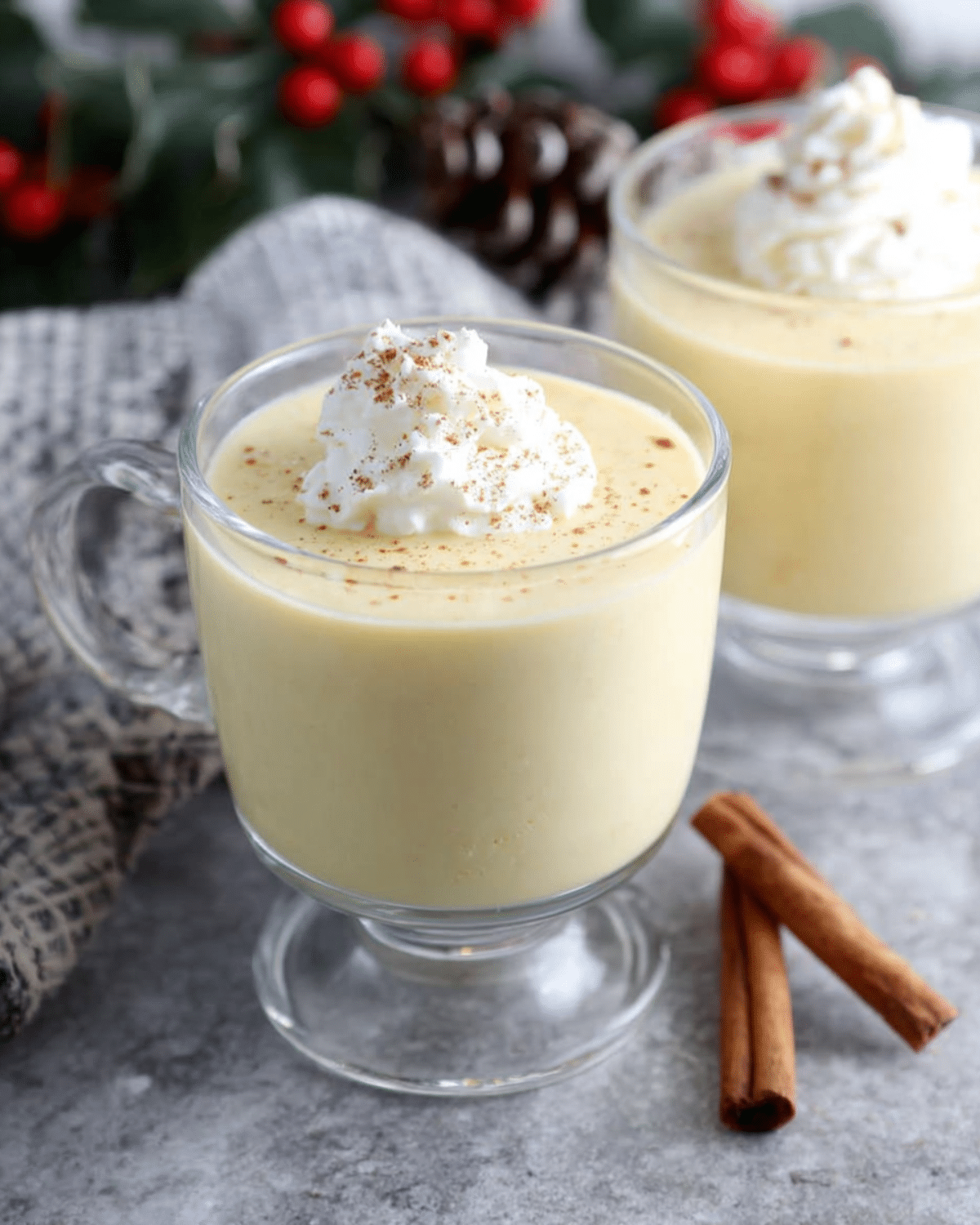 Christmas Nutmeg Vanilla Eggnog in a clear glass mug topped with whipped cream and sprinkled nutmeg, with cinnamon sticks on the side.