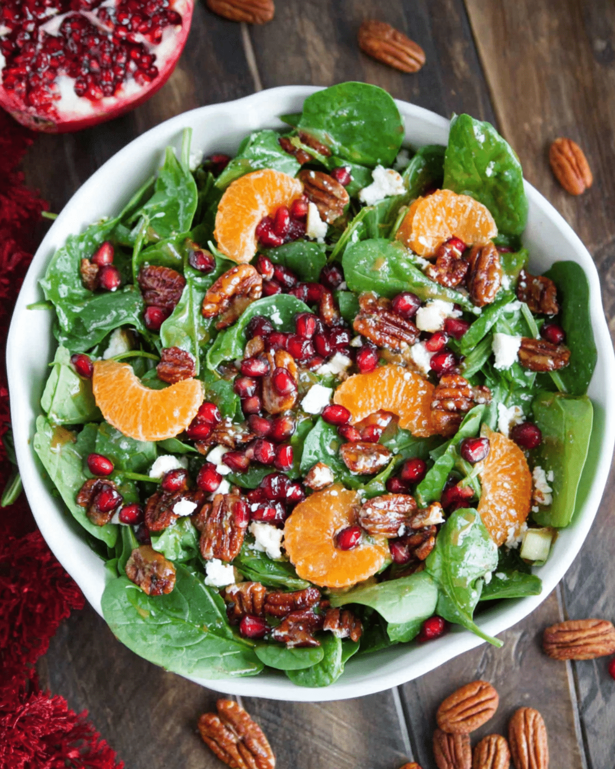 White bowl filled with Christmas salad made of spinach, mandarin orange slices, pomegranate seeds, candied pecans, and crumbled cheese, drizzled with honey mustard dressing.