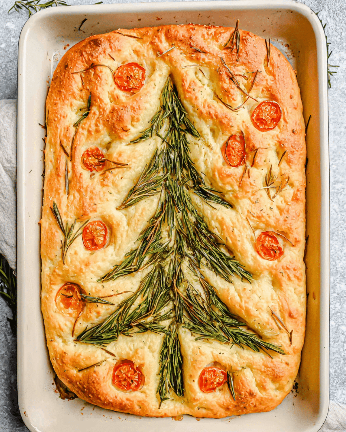 Rectangular focaccia bread decorated with rosemary sprigs arranged as a Christmas tree and cherry tomato ornaments.