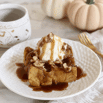 Pumpkin bread pudding topped with vanilla ice cream, pecans, and drizzled with brown sugar sauce on a white plate.