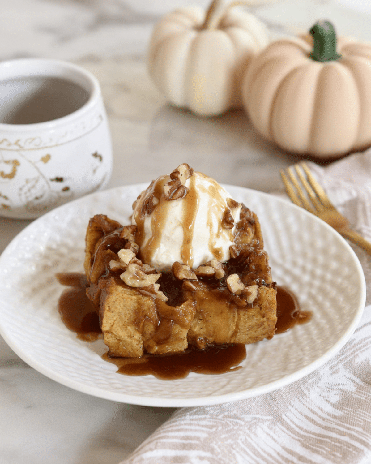 Pumpkin bread pudding topped with vanilla ice cream, pecans, and drizzled with brown sugar sauce on a white plate.