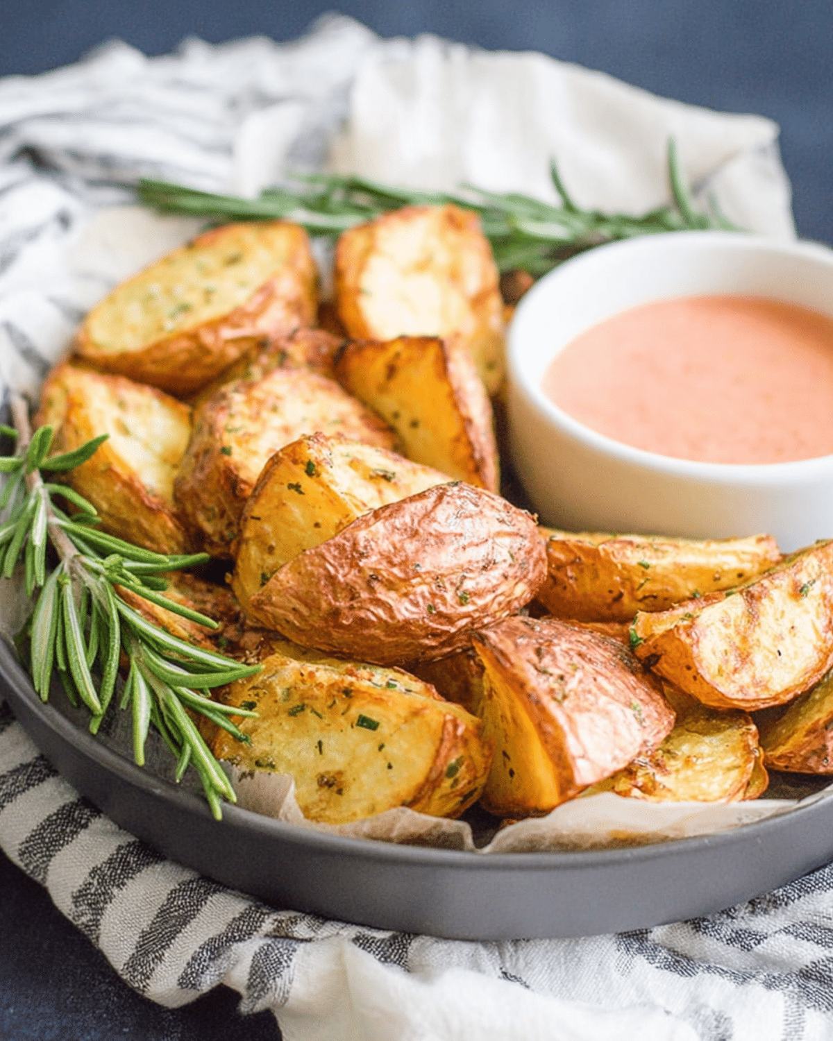 Crispy rosemary infused roast potatoes served in a gray dish with fresh rosemary sprigs and a bowl of dipping sauce.